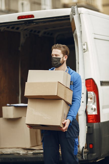 A male mover with light skin, wearing a black face mask, a blue jacket, and navy trousers, is standing at the back of a white moving van. He is holding three cardboard boxes of varying sizes, stacked